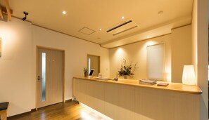 A reception area with a wooden reception desk, a potted plant, a flower arrangement, and a window with blinds.