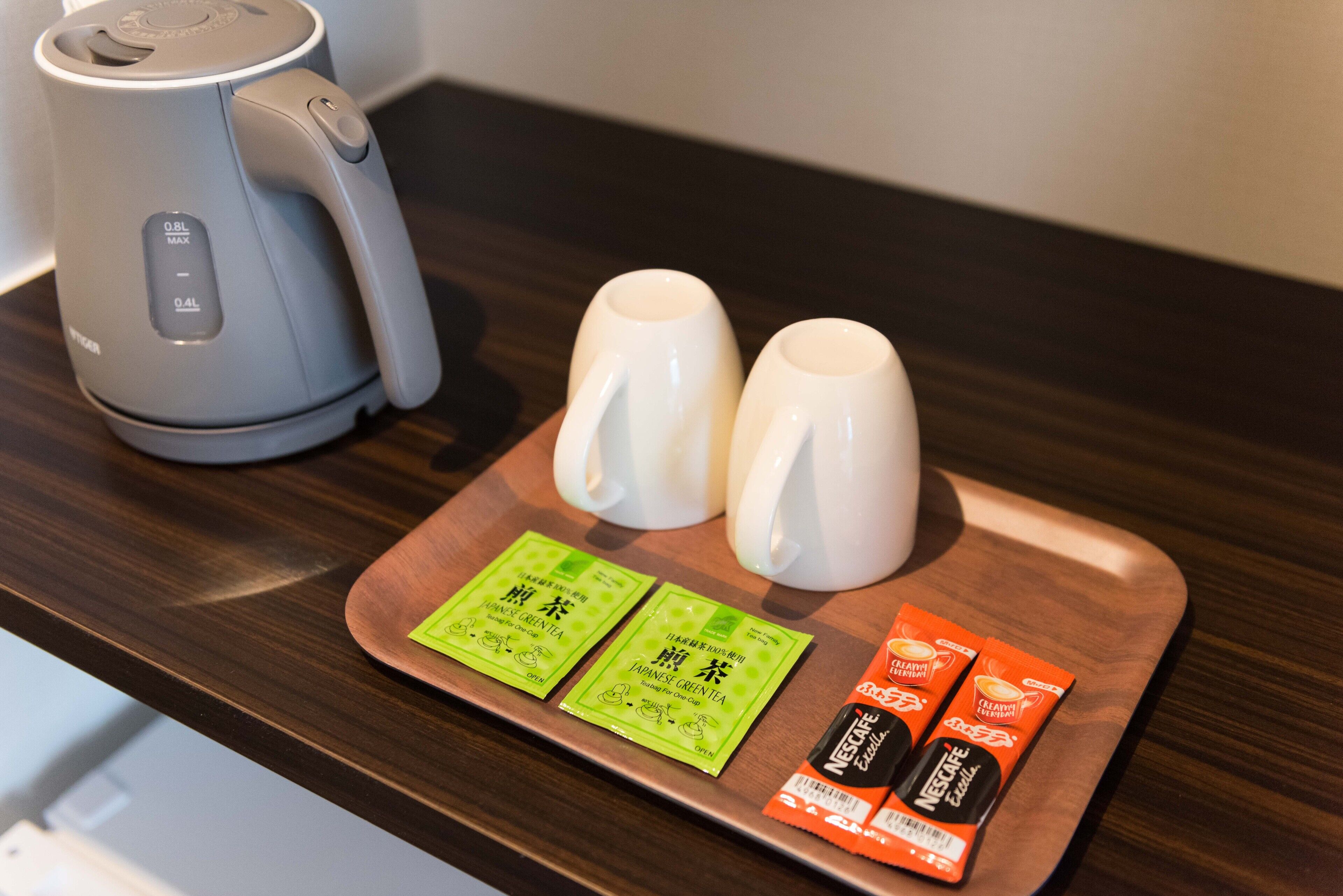 Una cafetera, dos tazas blancas y una bandeja con sobres de bebidas verdes y anaranjadas sobre una encimera de madera.