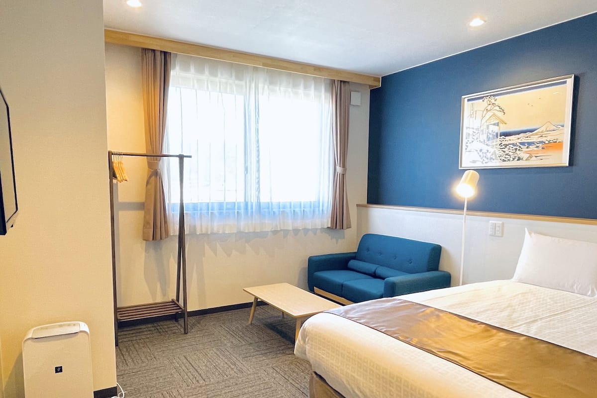 Quarto de hotel com uma cama, uma poltrona azul, uma mesinha, um aquecedor portátil e uma janela com cortinas.