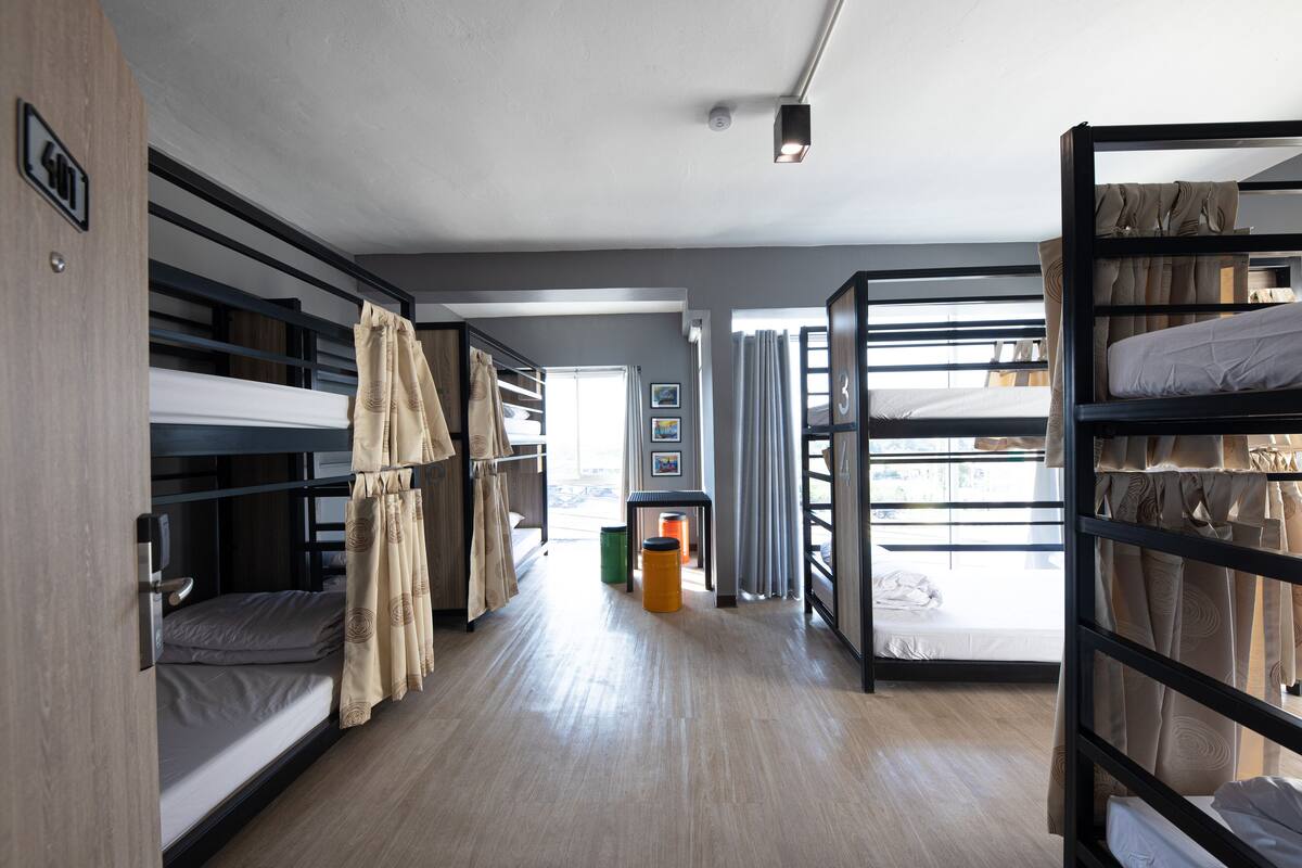 A room with bunk beds, a wooden floor, a trash bin, and a window with curtains.