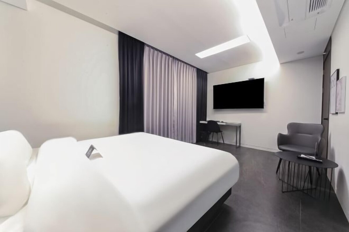 A modern hotel room with a large bed, a desk, a chair, and a TV mounted on the wall.