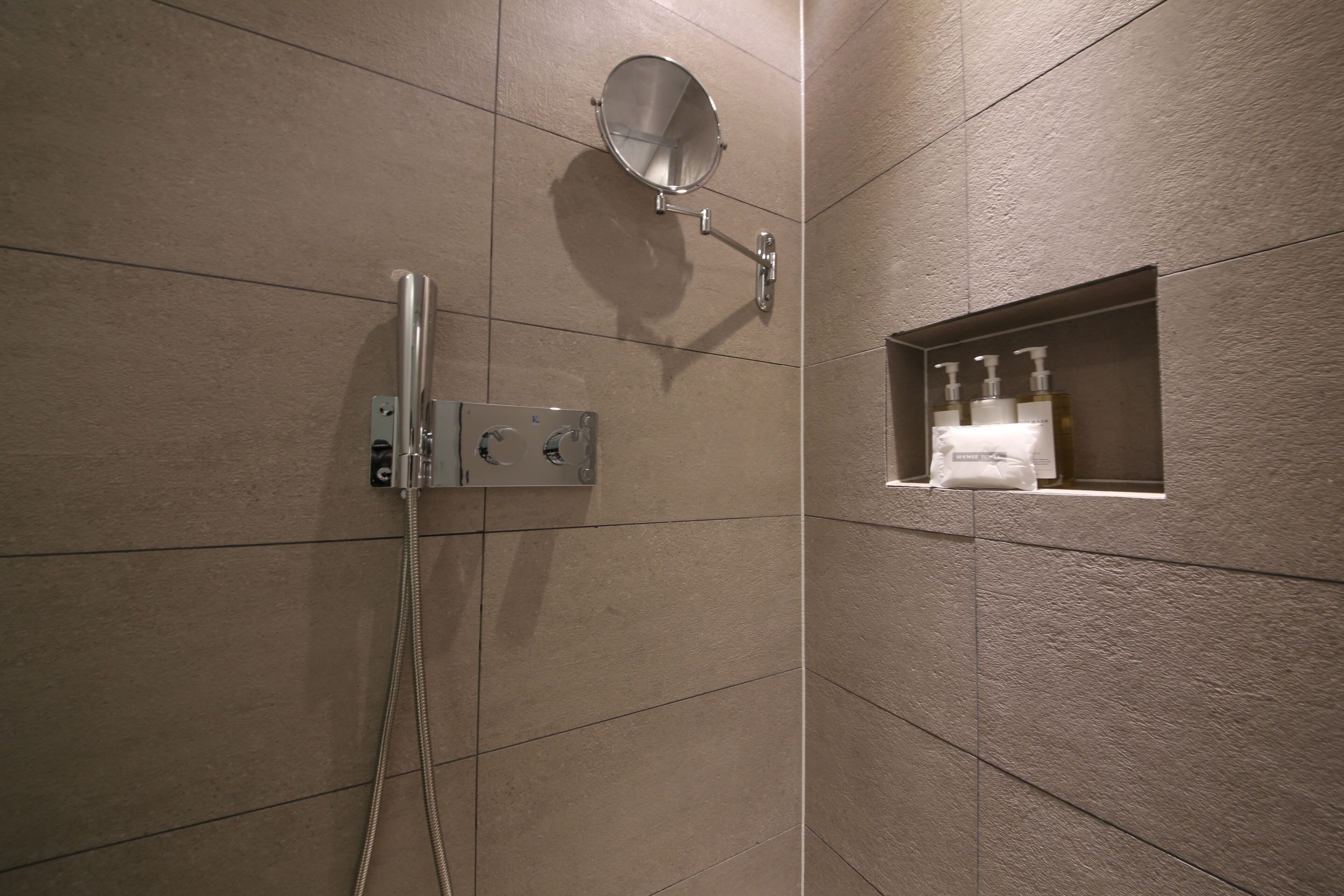 Un baño con ducha, cabezal de ducha manual, espejo montado en la pared y repisas con artículos de aseo.