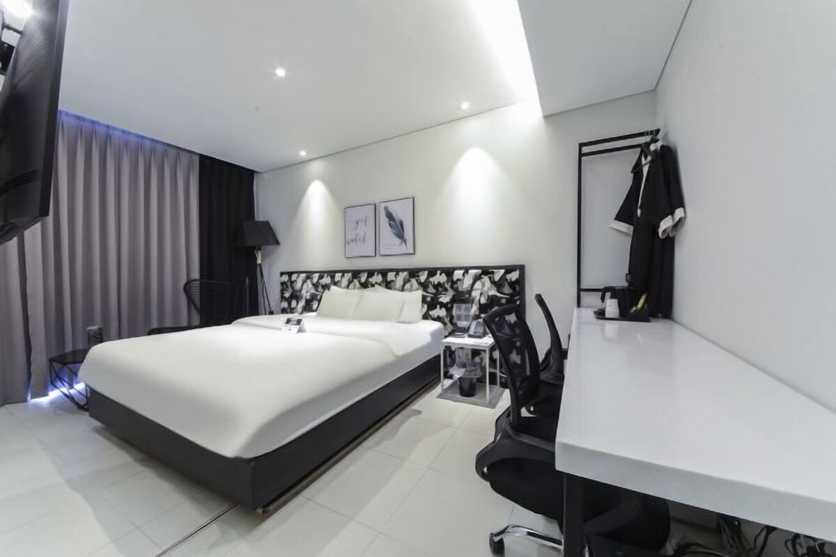 A modern hotel room with a large bed, bedside table, chair, and a desk with a chair.