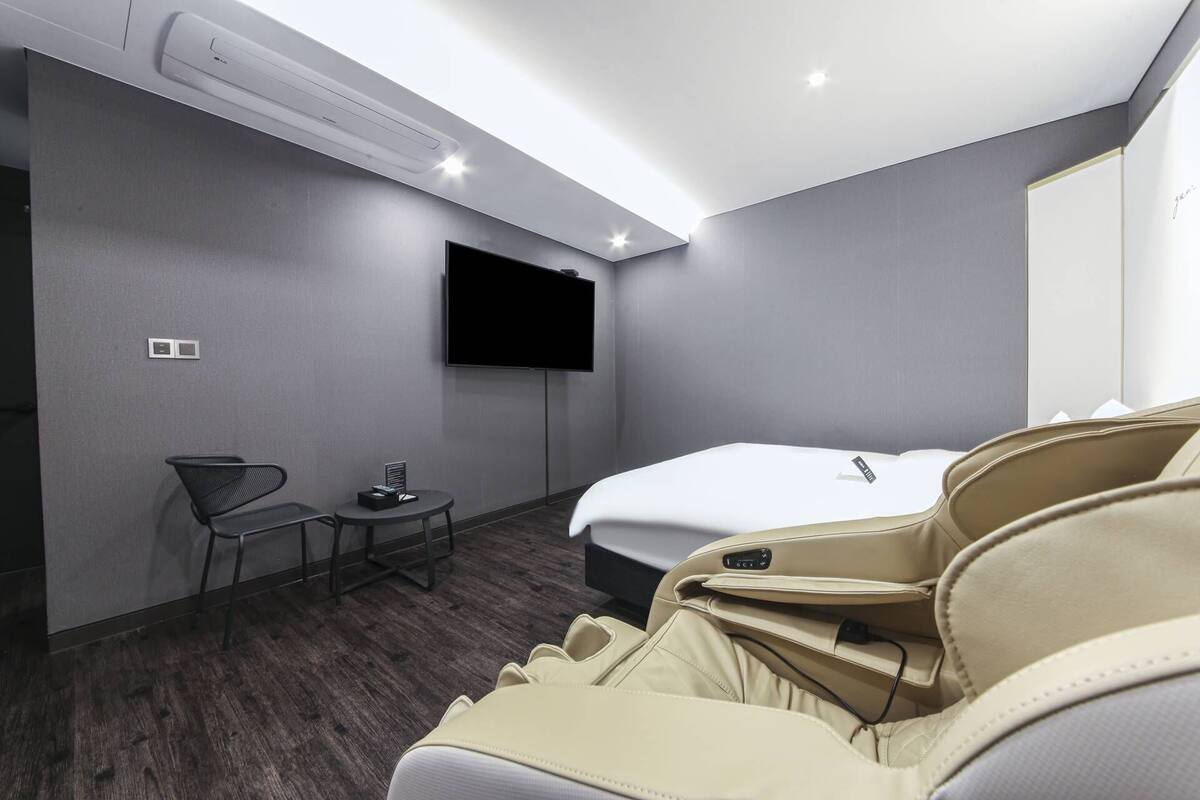 A hotel room with a bed, a TV mounted on the wall, a small table with a chair, and an air conditioning unit.