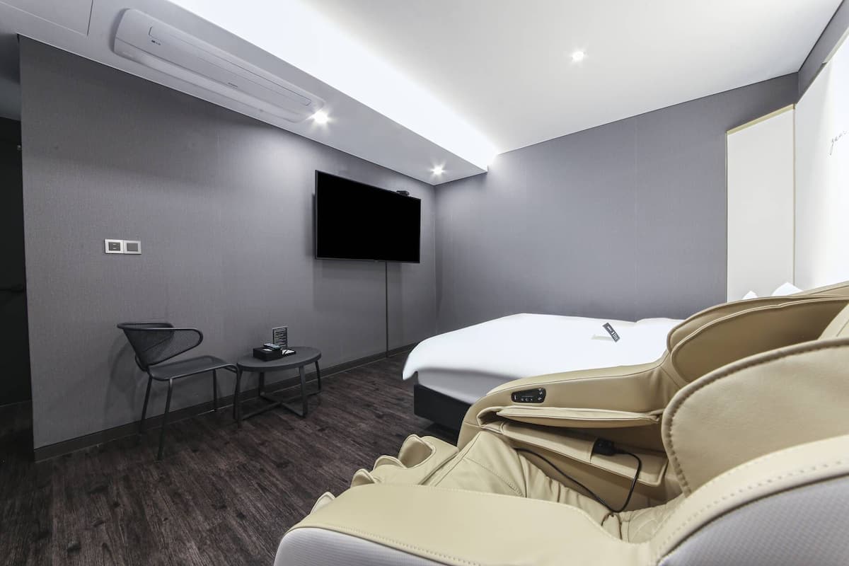 A hotel room with a bed, a TV mounted on the wall, a small table with a chair, and an air conditioning unit.