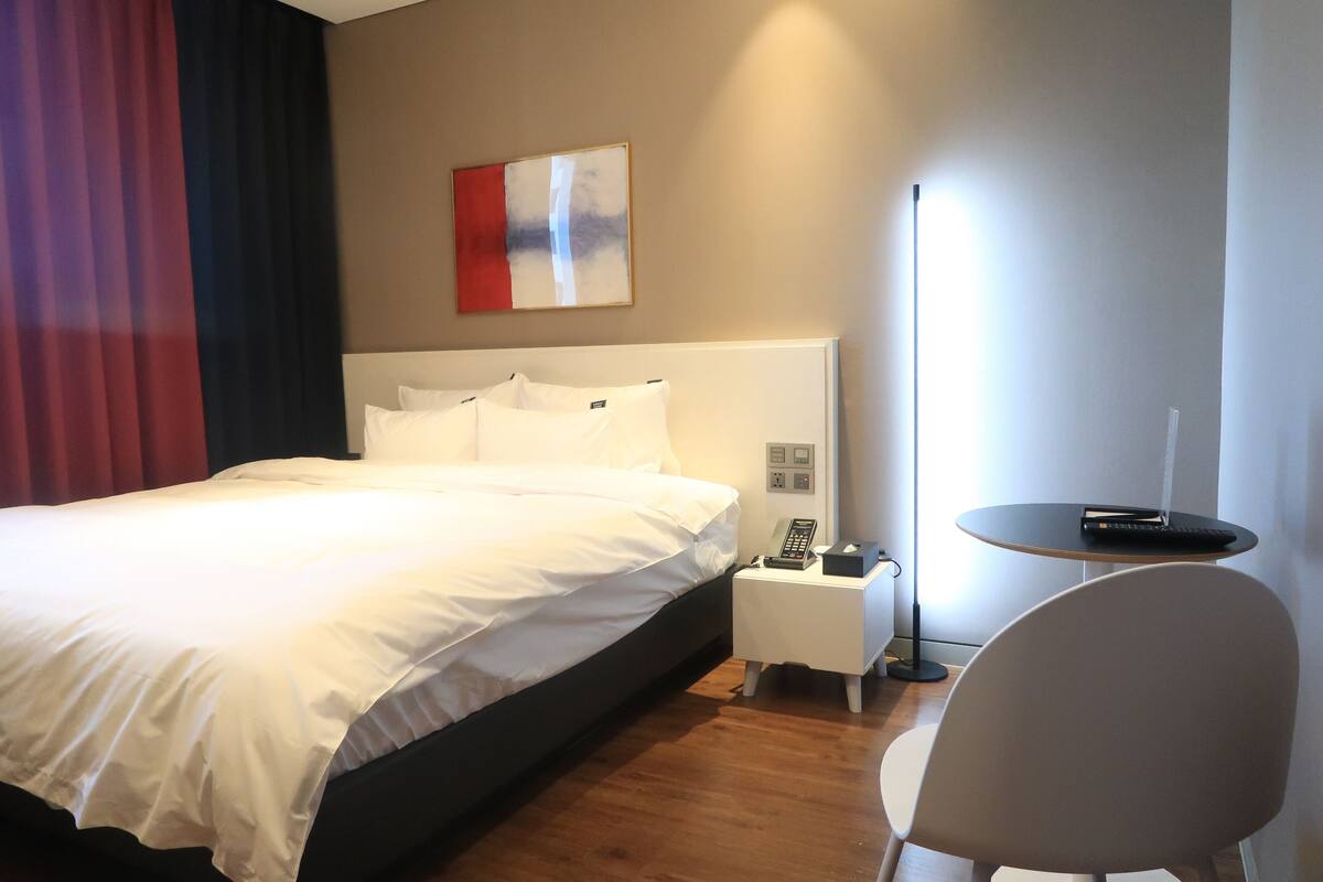 A hotel room with a large bed, bedside table with a phone and remote, a small table with a remote, and a chair.