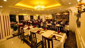 A large dining area with multiple tables and chairs, a chandelier, and a stone wall with framed pictures. - Le Bordeaux Sapa Hotel (Sa Pa)