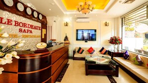A hotel lobby with a reception desk, a sofa, a coffee table, and a television displaying a cityscape. - Le Bordeaux Sapa Hotel (Sa Pa)