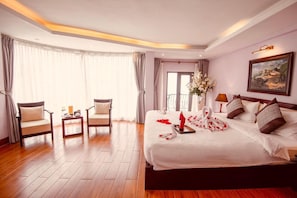Een hotelkamer met een groot bed, twee fauteuils, een tafeltje en een balkon met gordijnen.