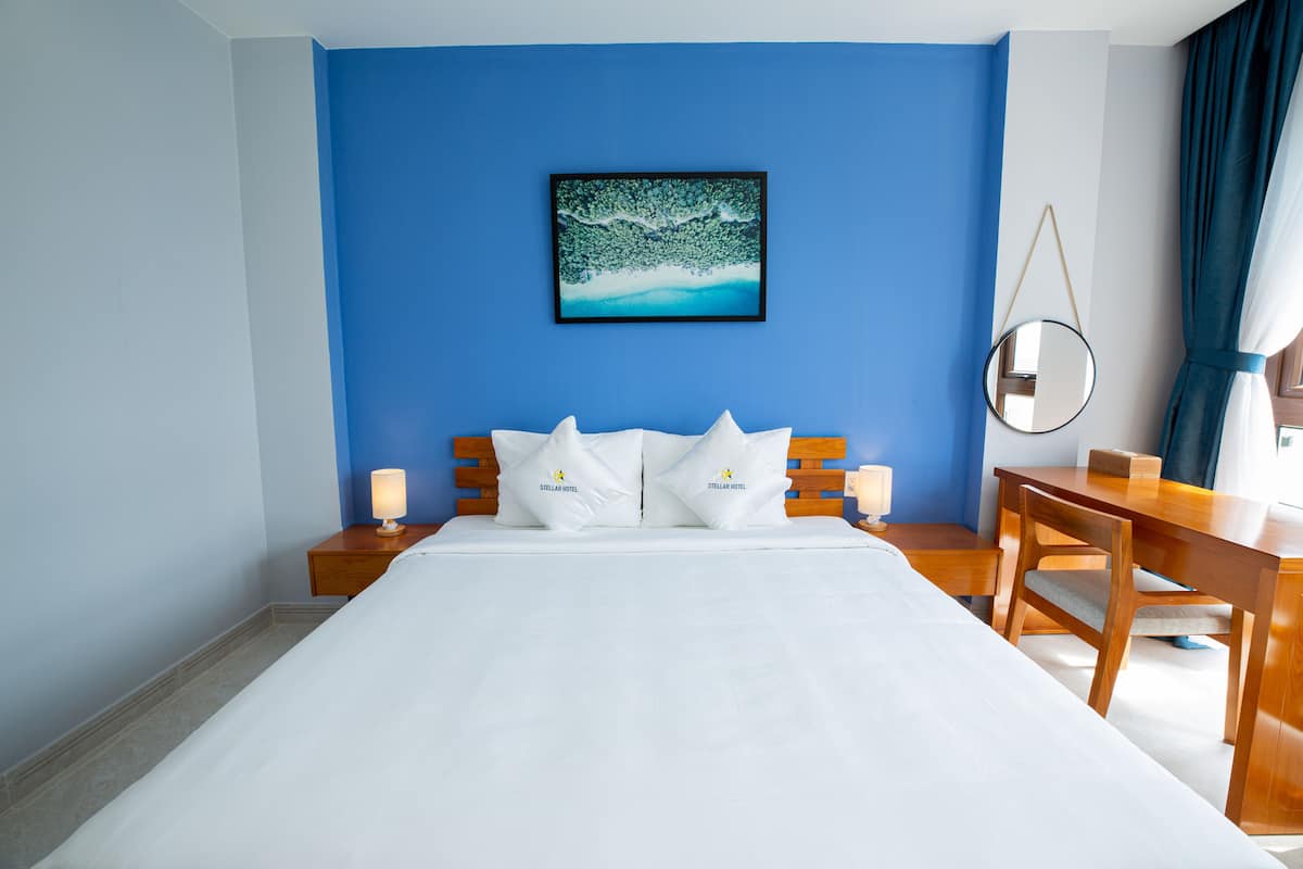 Habitación de hotel con una cama grande, una mesita de noche de madera con una lámpara, un escritorio con una silla y un cuadro en la pared.