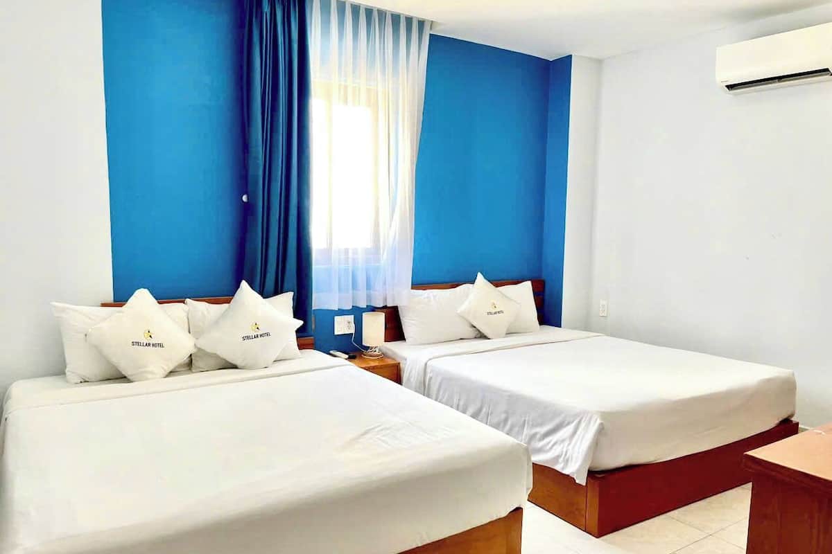 Habitación de hotel con dos camas individuales, una mesita de noche, un banco de madera y una ventana con cortinas traslúcidas.