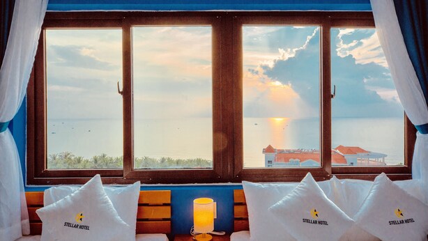 A room with a large window overlooking a beach, two beds with pillows labeled "Stellar Hotel", and a table with a lamp. - Stellar Hotel (Phu Quoc)