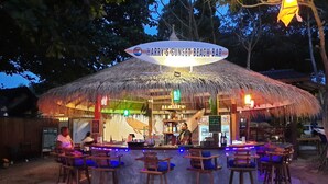 Un bar de playa llamado Harry's Sunset Beach Bar con techo de paja, taburetes de madera y un cartel que indica que sirve platos de San Salvador.