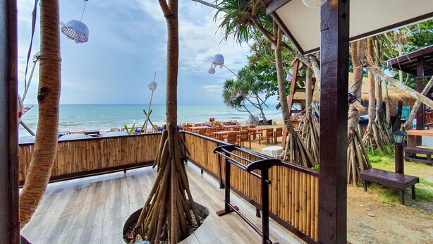 Terraza de madera con vista a la playa, con mesas y sillas, palmeras y vista al océano.