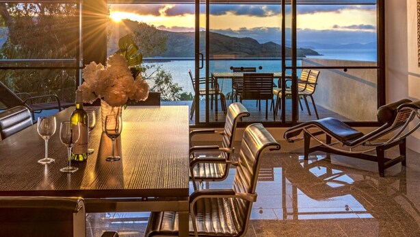 A dining area with a table set for two, a vase with flowers, and a view of the sea and hills. - Waves 3 Luxury 3 Bedroom (Whitsundays)