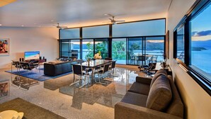 A modern living room with a large dining table, comfortable seating, and a view of the water. - Waves 3 Luxury 3 Bedroom (Whitsundays)