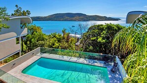 A swimming pool with a glass edge, surrounded by lush greenery and overlooking a coastal landscape with hills and a body of water. - Waves 3 Luxury 3 Bedroom (Whitsundays)