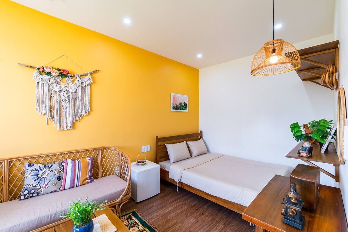 A compact room with a bed, a sofa, a small table, and a macrame wall hanging.