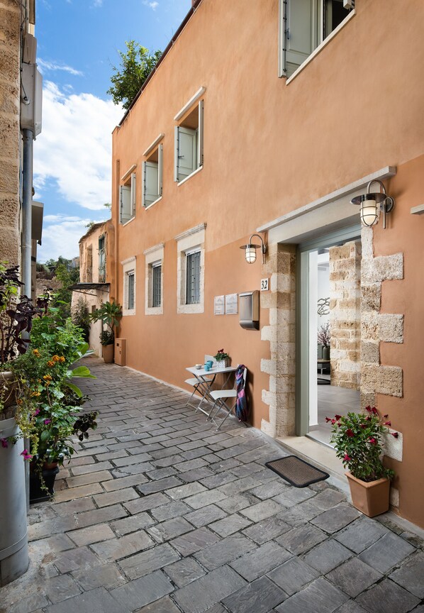A narrow cobblestone street with a building featuring a stone facade, a small table with chairs, and potted plants.