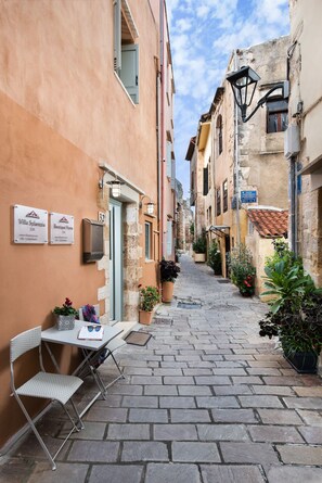 Una estrecha calle empedrada con una mesa y sillas frente a un edificio, con letreros que indican 'Villa Splanzia' y 'Boutique Home', y macetas con plantas a lo largo del camino.