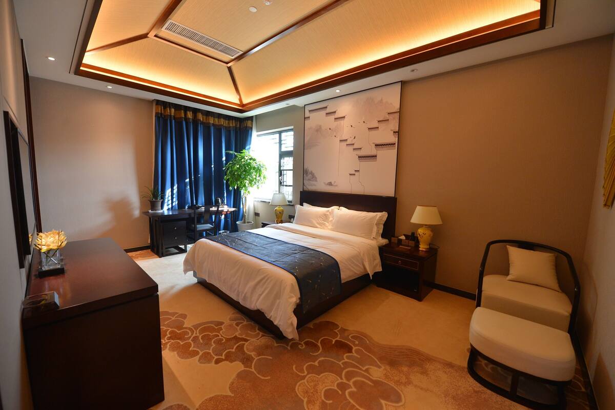 A modern hotel room with a large bed, bedside tables, a desk, a chair, and a large window with curtains.