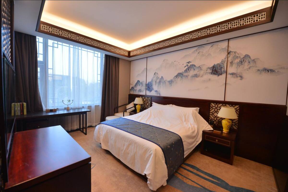 A hotel room with a large bed, a desk, a chair, a nightstand with a lamp, and a wall with a mountain painting.