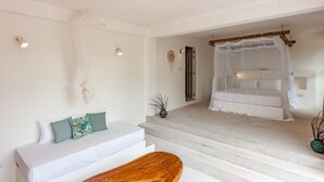 Un dormitorio con una cama blanca, un banco de madera, un sofá blanco con cojines verdes y una escalera que lleva a un balcón con dosel blanco.