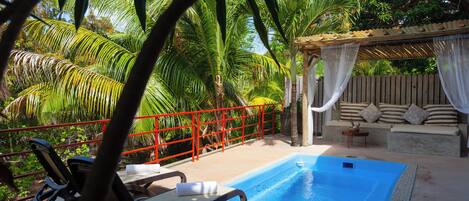 A pool area with a red railing, lounge chairs, and a covered seating area with cushions.