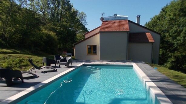Une piscine rectangulaire à l’eau d’un bleu limpide, entourée d’une terrasse en béton. On aperçoit des chaises longues et un petit bâtiment avec un toit recouvert de tuiles rouges à l’arrière-plan.