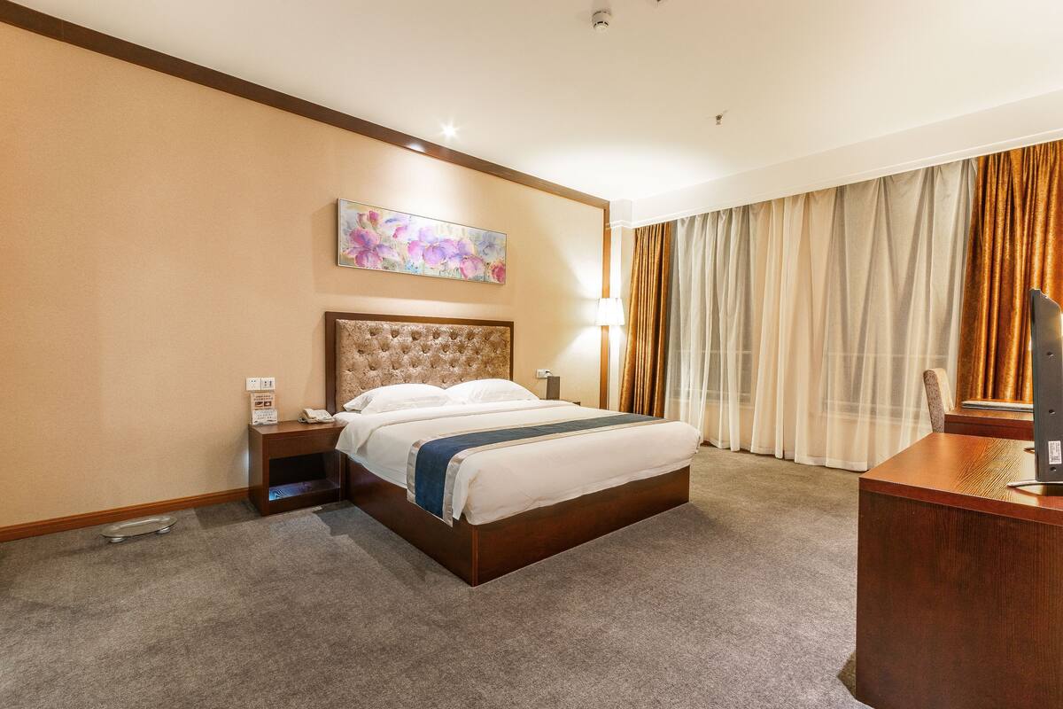Ein Hotelzimmer mit einem großen Bett, einem Holz-Kopfteil, einem Nachttisch, einem Schreibtisch und einem großen Fenster mit Vorhängen.