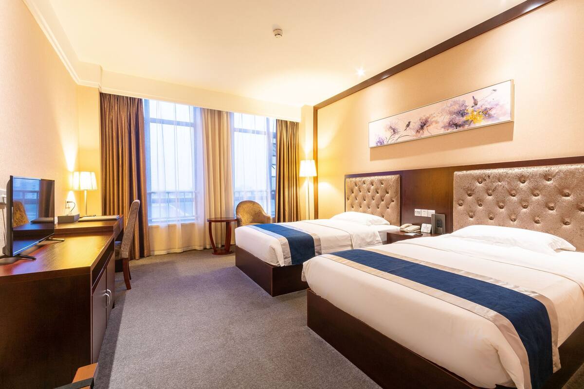 Ein Hotelzimmer mit zwei Betten, einem Schreibtisch, einem Sessel und einem großen Fenster mit Vorhängen.