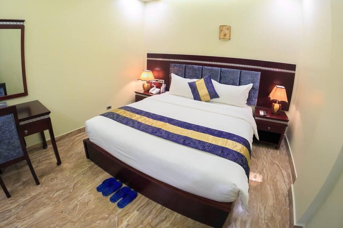 Uma cama bem arrumada com colcha listrada em branco e azul, duas luminárias de cabeceira, uma mesa de apoio, uma cadeira e um par de chinelos no chão.