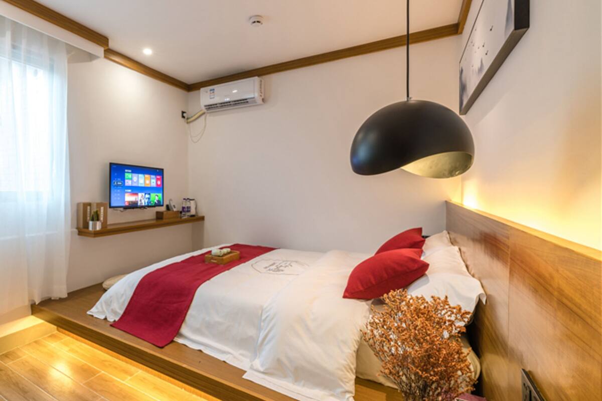 A hotel room with a bed, a TV, a shelf with items, and a hanging lamp.