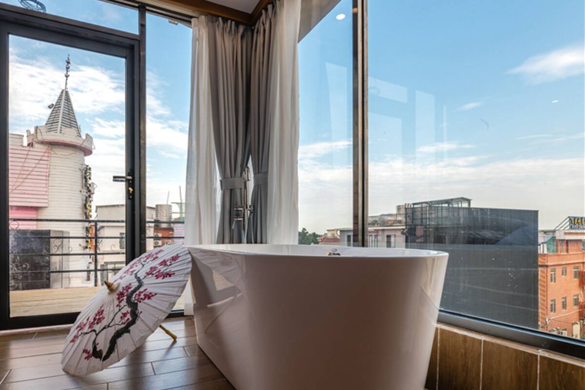 A modern bathroom with a large bathtub, a wooden bench, and a view of a cityscape through large windows.