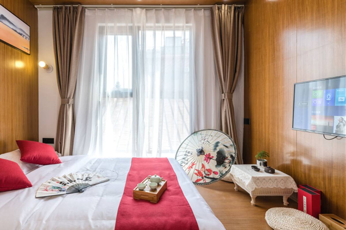 A hotel room with a large bed, red pillows, a fan, and a small table with a teapot and cups.