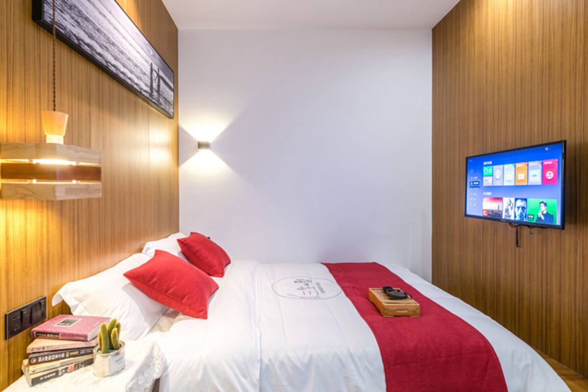 A modern hotel room with a bed, red pillows, a mounted TV displaying a home screen, and a wooden wall panel.