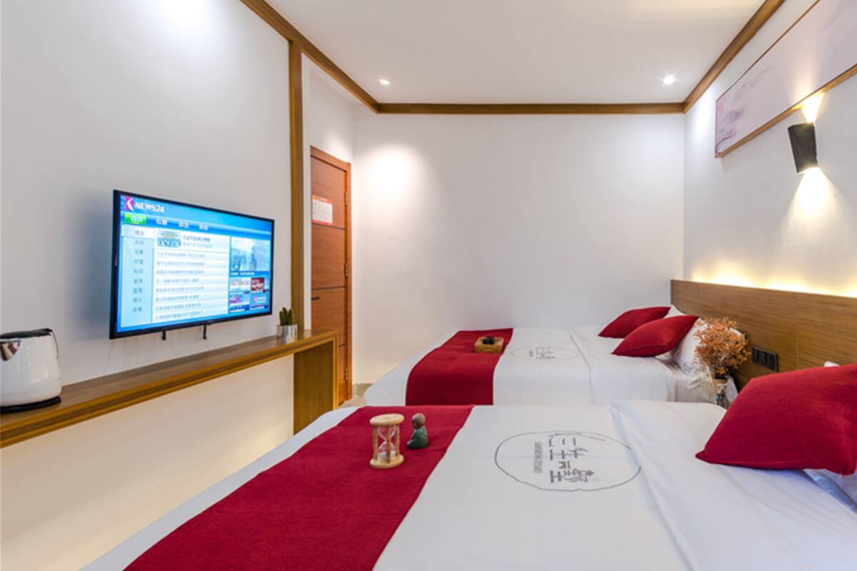 A hotel room with two beds, each with red pillows and a red runner. There is a television on the left wall displaying a news channel.