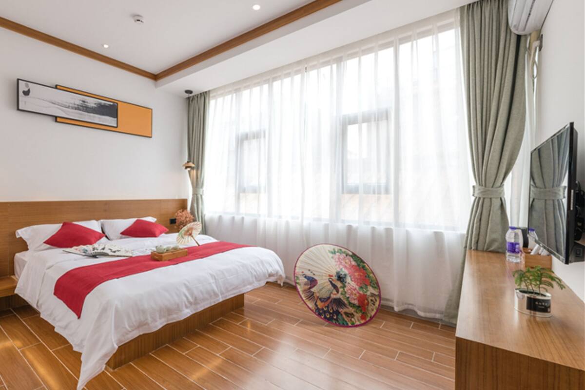 A modern hotel room with a wooden floor, a large bed with red and white bedding, a bedside table with books and a water bottle, a wall-mounted TV, and a large window with sheer curtains.