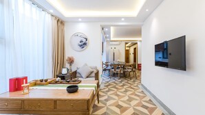 A modern hotel room with a wooden dining table, a sofa, a TV mounted on the wall, and a patterned floor.
