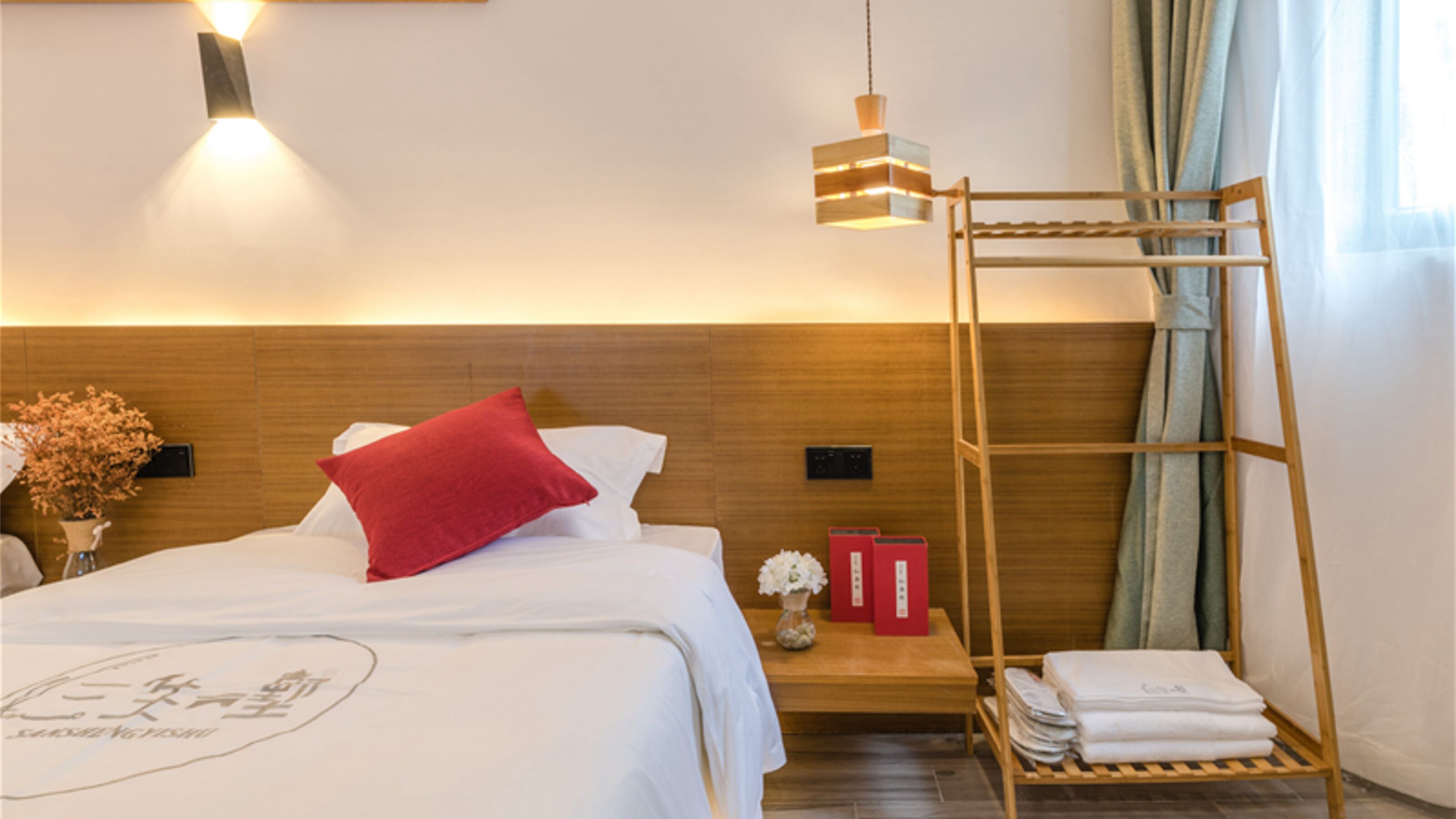 Ein Hotelzimmer mit einem Holz-Kopfteil, einem Bett mit weißer Bettwäsche und einem roten Kopfkissen, einem Nachttisch mit Blumen in einer Vase, einer Holzleiter und einer wandmontierten Leuchte.