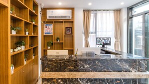 A hotel reception area with a marble counter, wooden shelving with plants, an air conditioning unit, a framed certificate, and a monitor displaying multiple images.