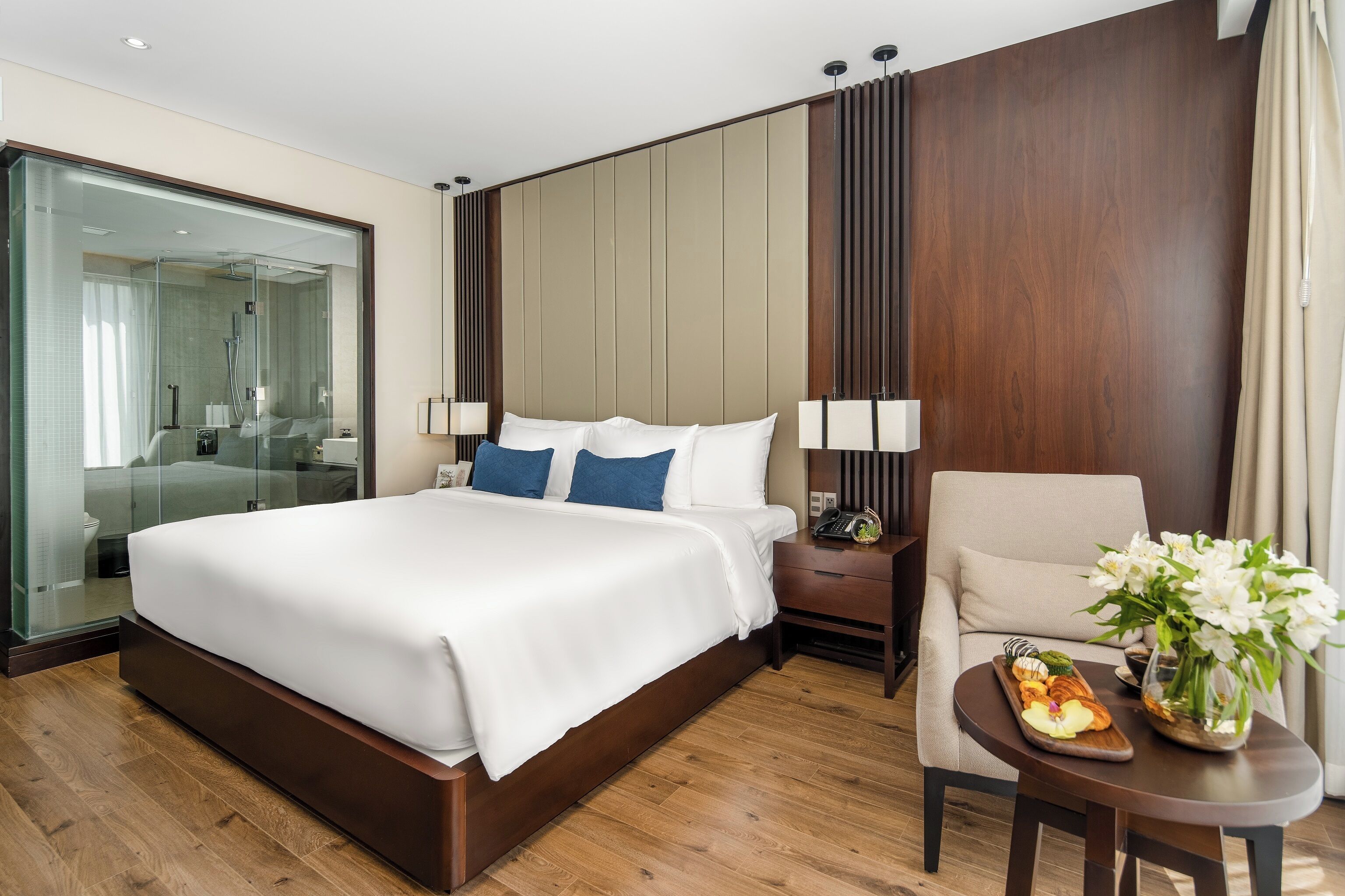 A modern hotel room with a large bed, a glass-enclosed shower, a wooden nightstand, a chair, and a small table with flowers and a tray of food.