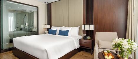 A modern hotel room with a large bed, a glass-enclosed shower, a wooden nightstand, a chair, and a small table with flowers and a tray of food.
