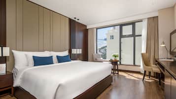 Una habitación de hotel moderna con una cama grande, un escritorio y vistas a un paisaje urbano a través de un amplio ventanal.