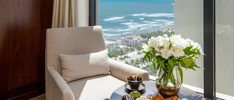 A room with a view of the ocean, a chair, a table with a tray of pastries, and a vase of flowers.