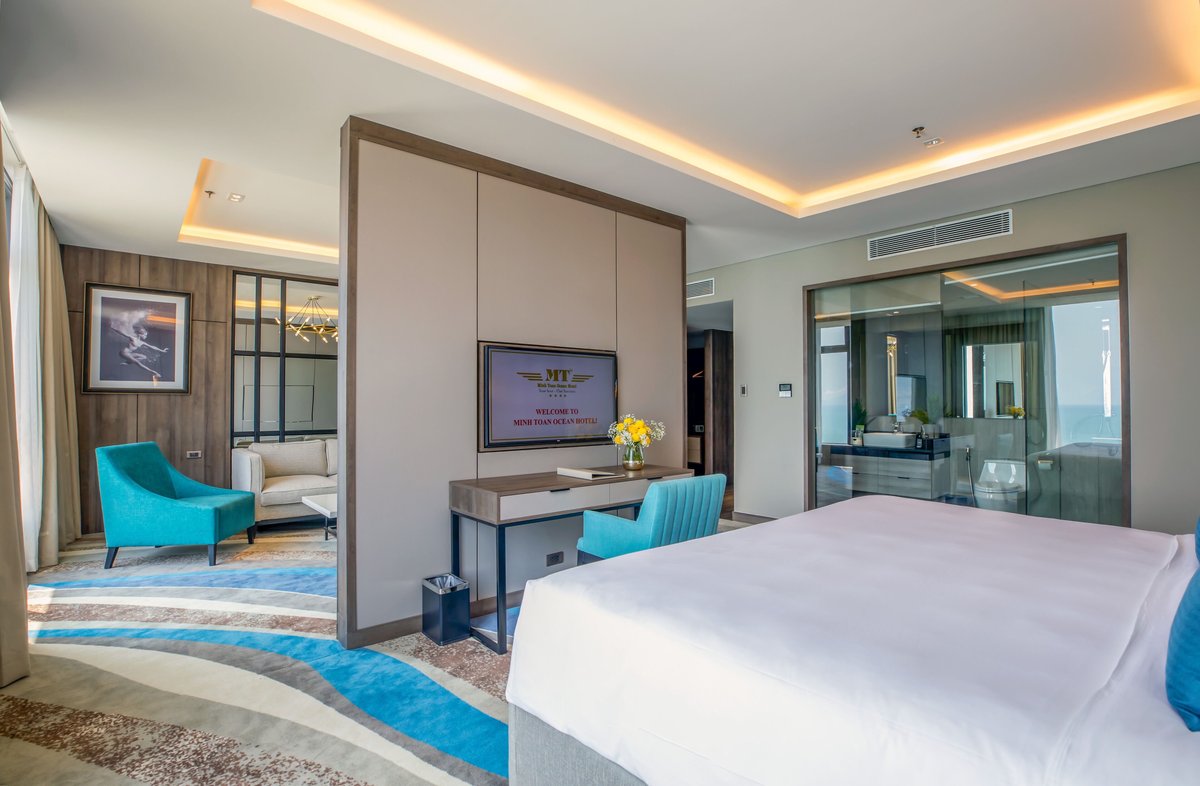 A hotel room with a large bed, a desk with a chair, a sofa, a TV, and a bathroom visible through a glass door.