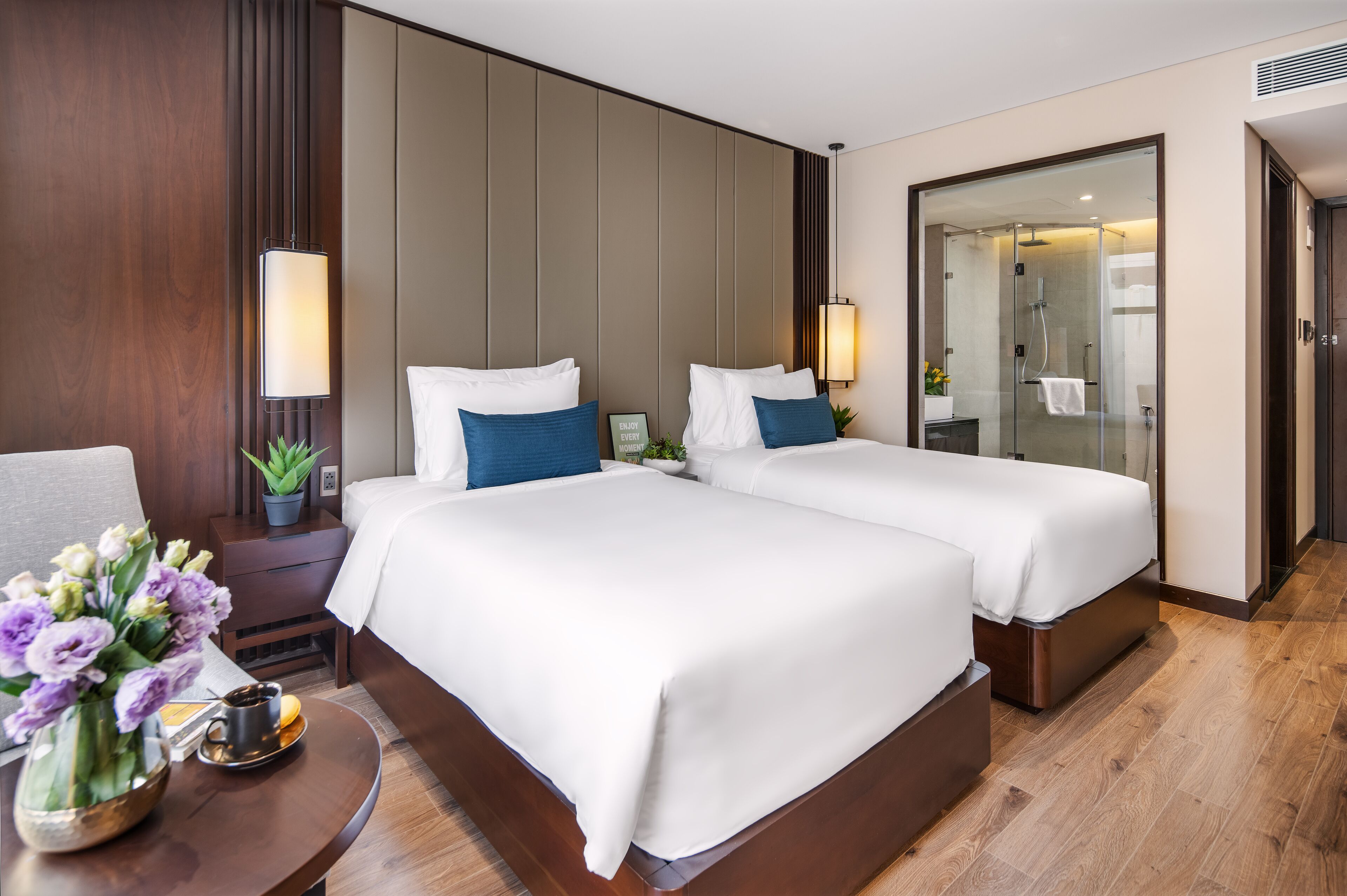 Een hotelkamer met twee bedden, een kleine tafel met een bloemenvaas, een nachtkastje en een douchecabine met glazen wanden.