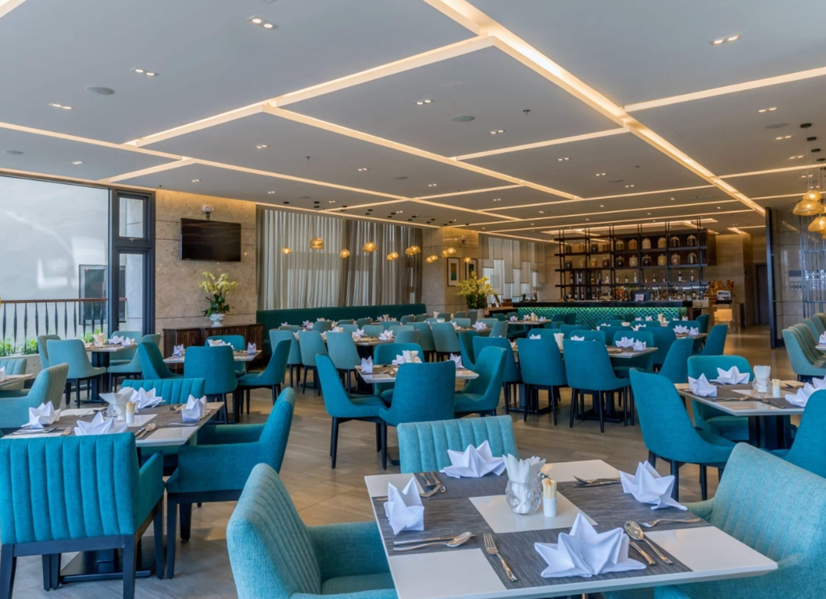 Un restaurante moderno con sillas y mesas color turquesa, una zona de bar y un amplio ventanal que proporciona luz natural.