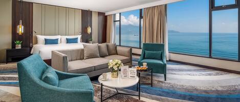 Habitación de hotel con cama, sofá, dos sillones, una mesita de centro y vista al mar.