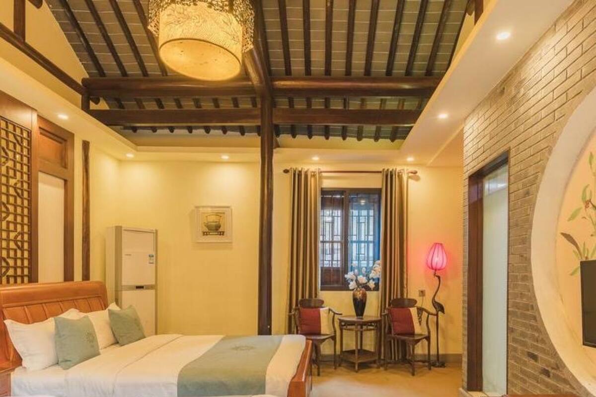 A hotel room with two beds, a wooden ceiling, a brick wall, a window with curtains, a small table with chairs, and a lamp.